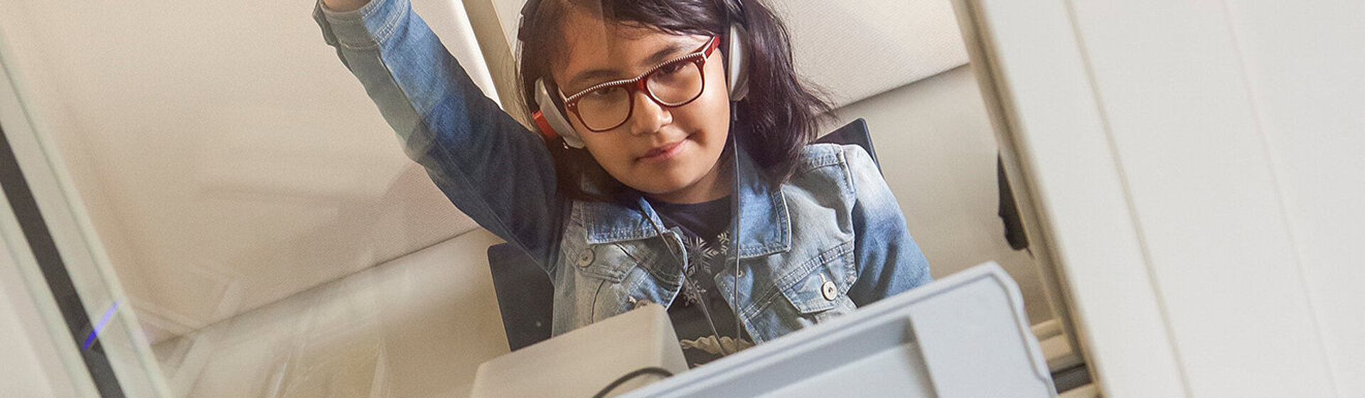 Ein Kind mit Kopfhörer auf den Ohren sitzt in einer Hörtestkabine und hebt den Arm