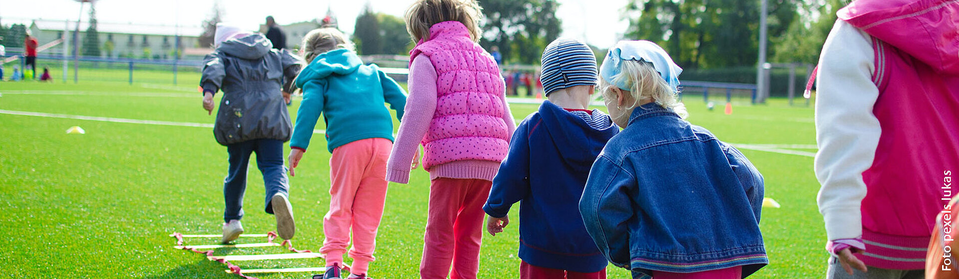 Kinder auf einer grünen Wiese in einer Reihe hintereinander machen ein Geschicklichkeits- und Hüpfspiel.