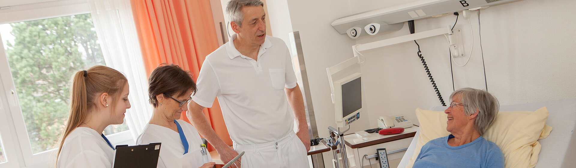 Prof. Dr. Michael Neipp und zwei medizinische Fachkräfte im Gespräch mit einer Patientin, die im Bett liegt. Prof. Dr. Michael Neipp und zwei medizinische Fachkräfte im Gespräch mit einer Patientin, die im Bett liegt.
