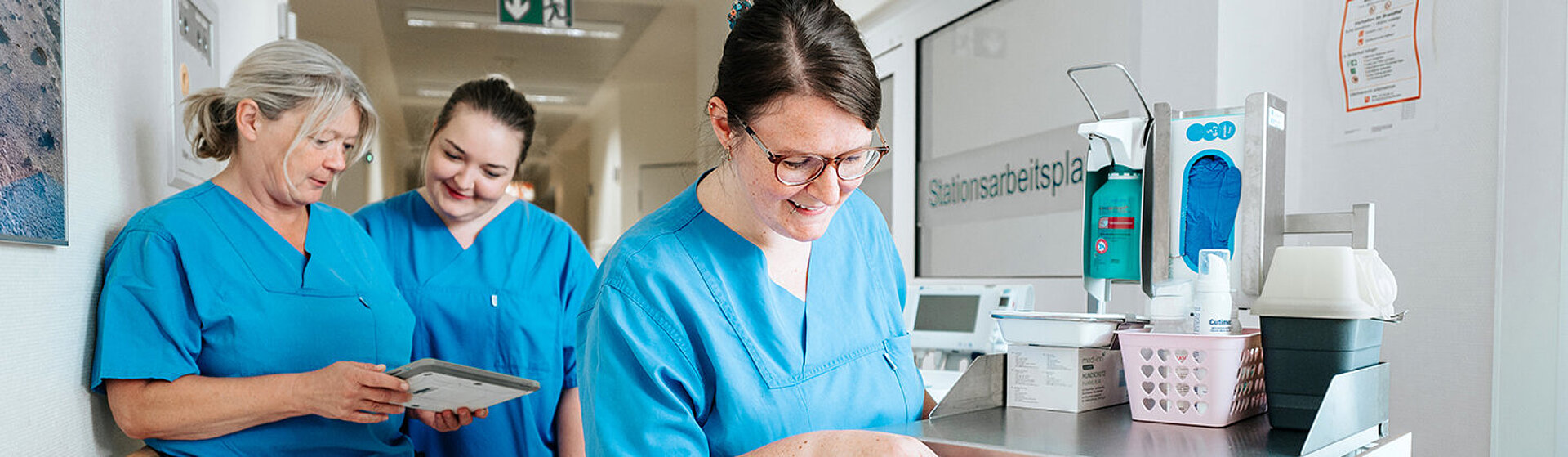 Drei Pflegefachkräfte stehen im Flur, zwei schauen auf ein Tablet, die dritte öffnet eine Schublade vom Medikamentenwagen.
