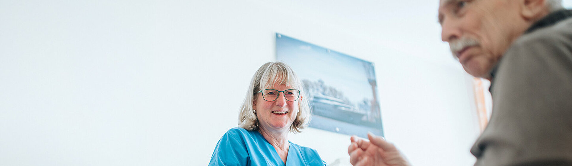 Patient sitzt im Rollstuhl, Pflegefachkraft lächelt ihn an und und gibt ihm einen Gehwagen. 