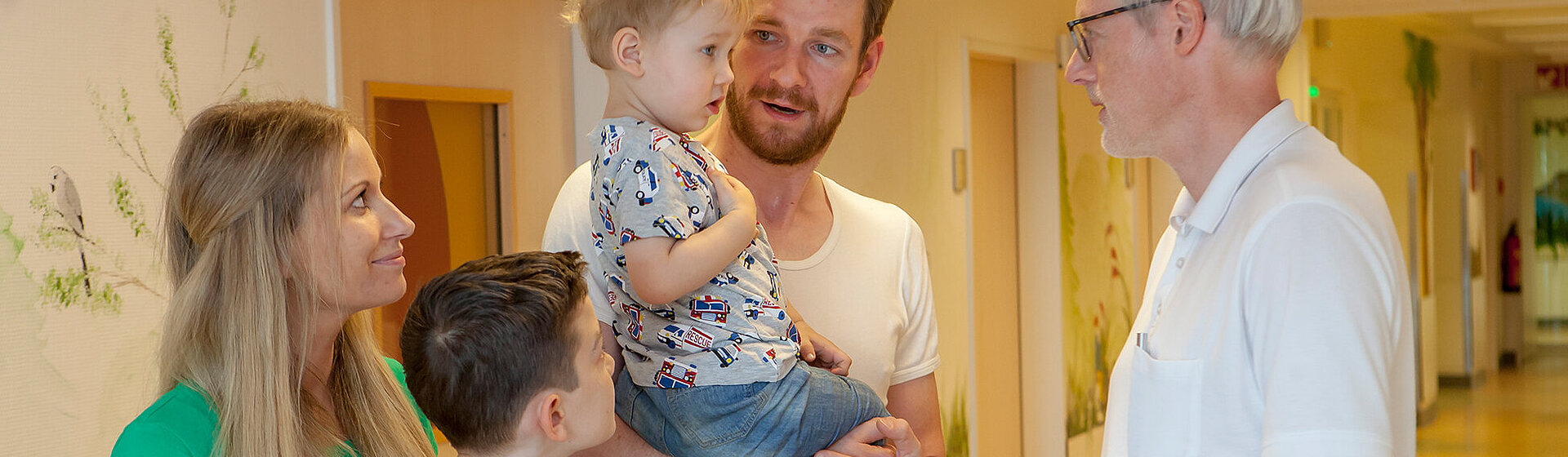 Chefarzt Dr. med. Georg Hillebrand (rechts) unterhält sich mit einem kleinen Patienten, der auf dem Arm des Vaters zusammen mit seiner Mutter und dem großen Bruder im Flur der Kinderklinik steht. Chefarzt Dr. med. Georg Hillebrand (rechts) unterhält sich mit einem kleinen Patienten, der auf dem Arm des Vaters zusammen mit seiner Mutter und dem großen Bruder im Flur der Kinderklinik steht.