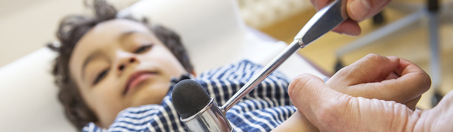 Ein Kind liegt auf einer Untersuchungsliege und wir von einem Arzt mit dem Reflexhammer untersucht. Ein Kind liegt auf einer Untersuchungsliege und wir von einem Arzt mit dem Reflexhammer untersucht.