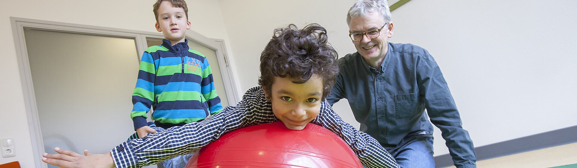 Physiotherapeut Sven Olde mit zwei Kindern in der Therapiesituation auf einer Gymnastikmatte, ein Kind liegt bäuchlings auf einem roten Gymnastikball, das zweite Kind hält die Füße des ersten Kindes. Physiotherapeut Sven Olde mit zwei Kindern in der Therapiesituation auf einer Gymnastikmatte, ein Kind liegt bäuchlings auf einem roten Gymnastikball, das zweite Kind hält die Füße des ersten Kindes.