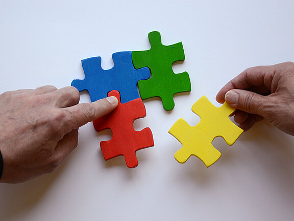 Es sind zwei Hände und vier bunte Puzzleteile abgebildet