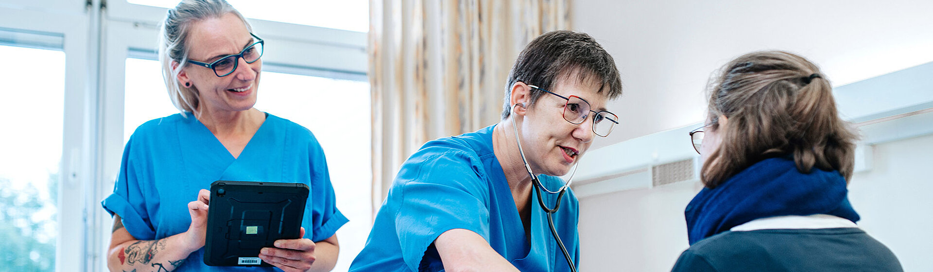 Eine Pflegefachkraft misst Blutdruck, die anderen hält ein Tablet in der Hand.