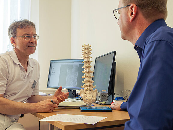 Dr. med. Wolfram Kluge klärt einen Patienten anhand eines Wirbelsäulenmodells über die SCS-Rückemankstimulation auf.