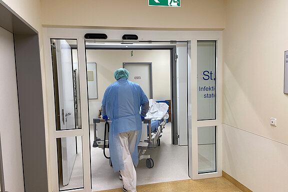 Ein Mitarbeiter mit Schutzkleidung schiebt ein PatientInnenbett durch die automatische Tür der Station 6.