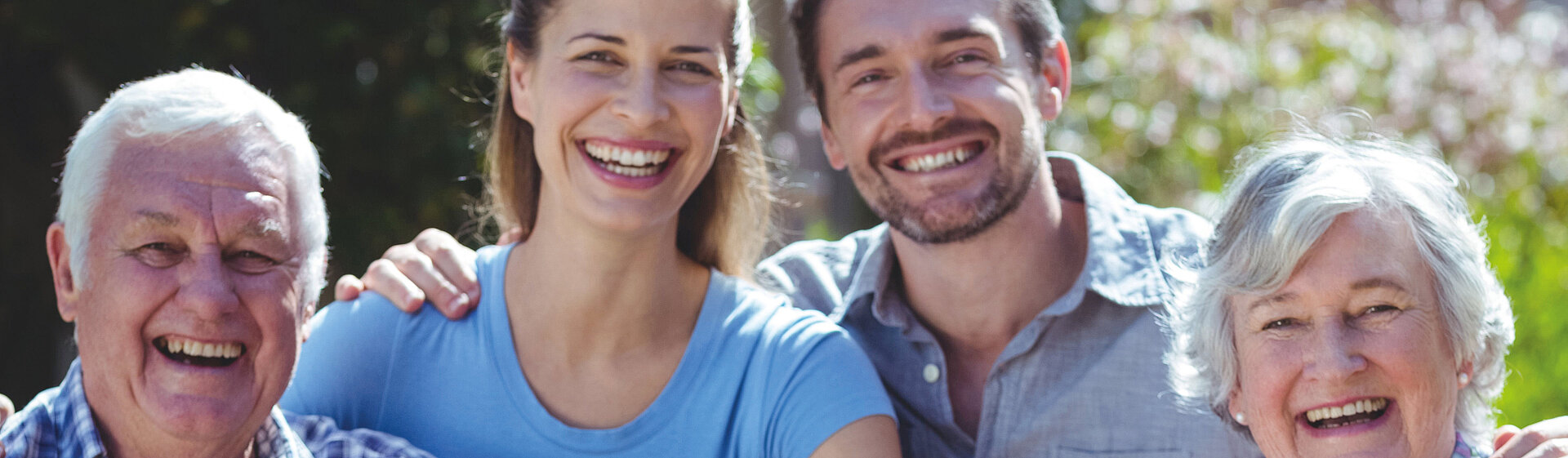 Eine fröhliche Familie bestehend aus Großeltern, Eltern und Kindern sitzen zusammen im Grünen und lächeln in die Kamera.
