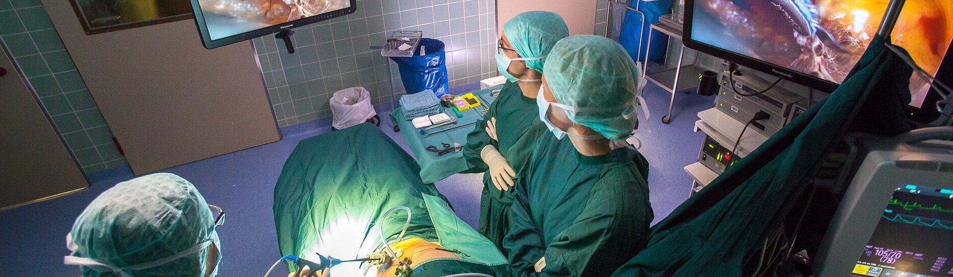 Minimalinvasive Operation im OP-Saal, drei medizinische Fachkräfte stehen am OP-Tisch und blicken auf den OP-Monitor.