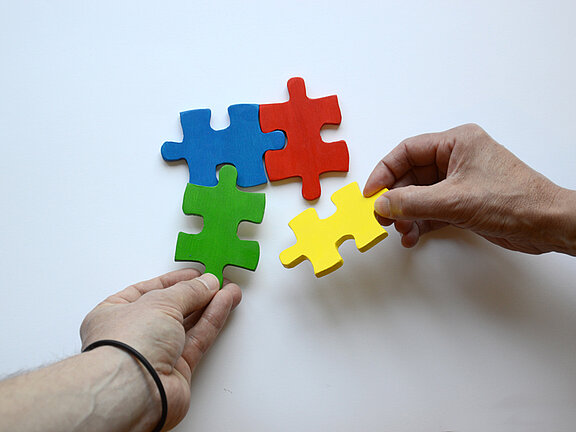 Es sind zwei Hände und vier bunte Puzzleteile abgebildet