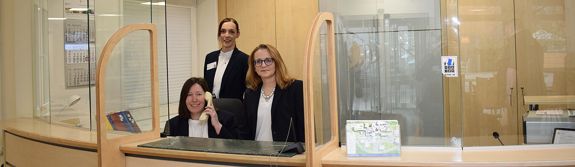 Drei Rezeptionsmitarbeiterinnen lächeln freundlich, eine telefoniert. Drei Rezeptionsmitarbeiterinnen lächeln freundlich, eine telefoniert.