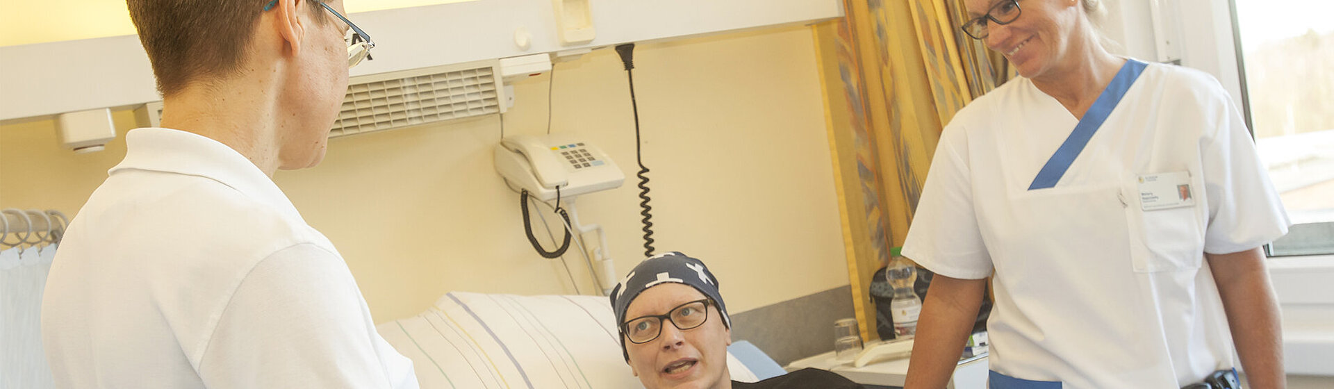 Patientin liegt im Bett, zwei Pflegefachkräfte stehen neben dem Bett und lächeln sie an. Patientin liegt im Bett, zwei Pflegefachkräfte stehen neben dem Bett und lächeln sie an.