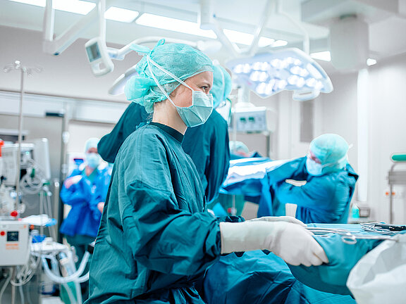 Arbeitssituation in einem Operationssaal, im Vordergrund eine medizinische Fachkraft in OP-Kleidung, OP-Maske und Handschuhen mit OP-Besteck